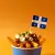A serving of poutine in a blue paper cup, topped with cheese curds, gravy, and chopped parsley, with a small Quebec flag on a toothpick placed in the center. The background is a solid orange color.