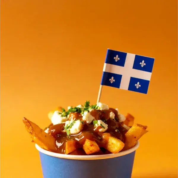 A serving of poutine in a blue paper cup, topped with cheese curds, gravy, and chopped parsley, with a small Quebec flag on a toothpick placed in the center. The background is a solid orange color.